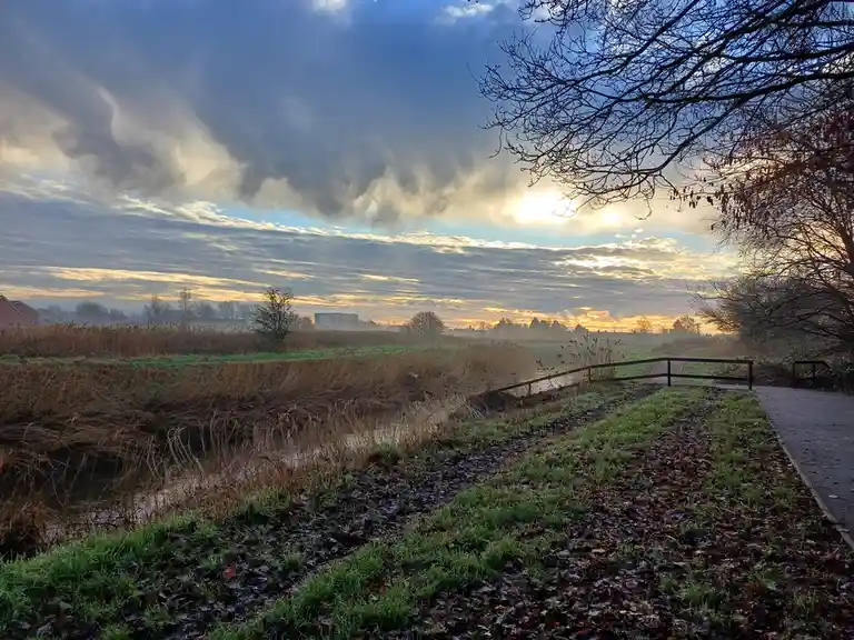 A scenic outdoor photo shows a misty, early morning landscape with a path alongside a canal, bare trees, and a dramatic…