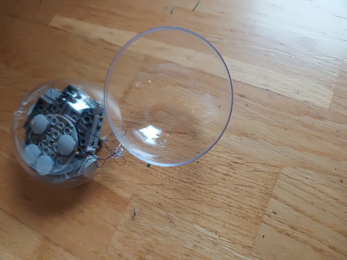 A clear plastic ball with a gray LEGO-like robotic mechanism inside sits on a wooden floor next to its empty transparent…