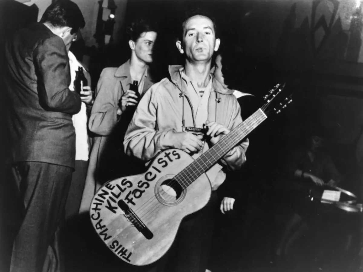 A black-and-white photo shows a man holding an acoustic guitar with the words 'THIS MACHINE KILLS FASCISTS' written on its…