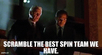 The image shows a still from a movie featuring two men in suits sitting at a table. The man on the left, who appears older…