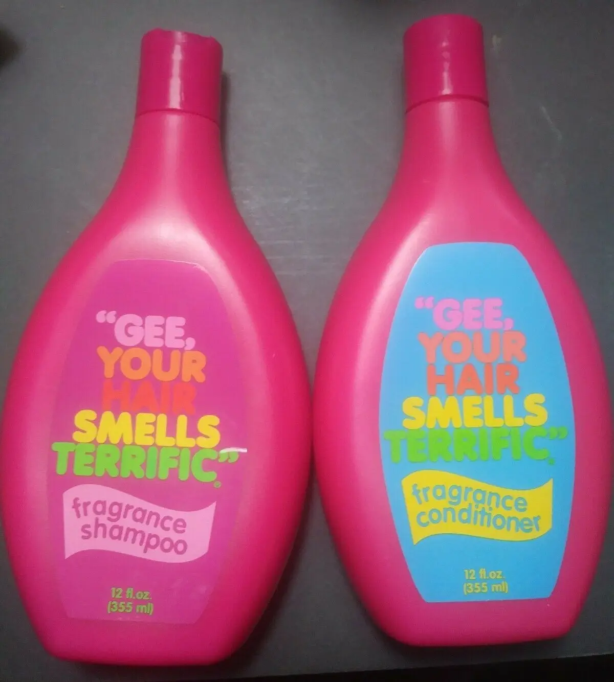 Two bright pink bottles of hair care products sit side by side on a dark surface. The left bottle, labeled 'fragrance…