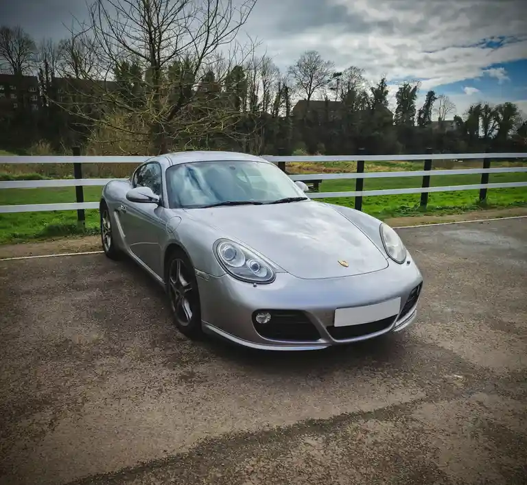 A silver Porsche Boxster (model 987) is parked on a paved surface next to a white fence, with grass and trees in the…