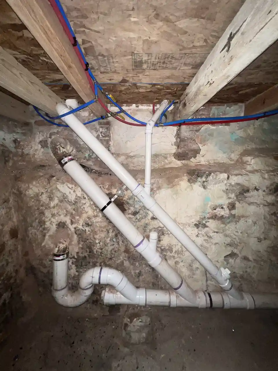 A photo shows a chaotic tangle of white PVC pipes and red/blue wires running across a rough, unfinished basement ceiling and…
