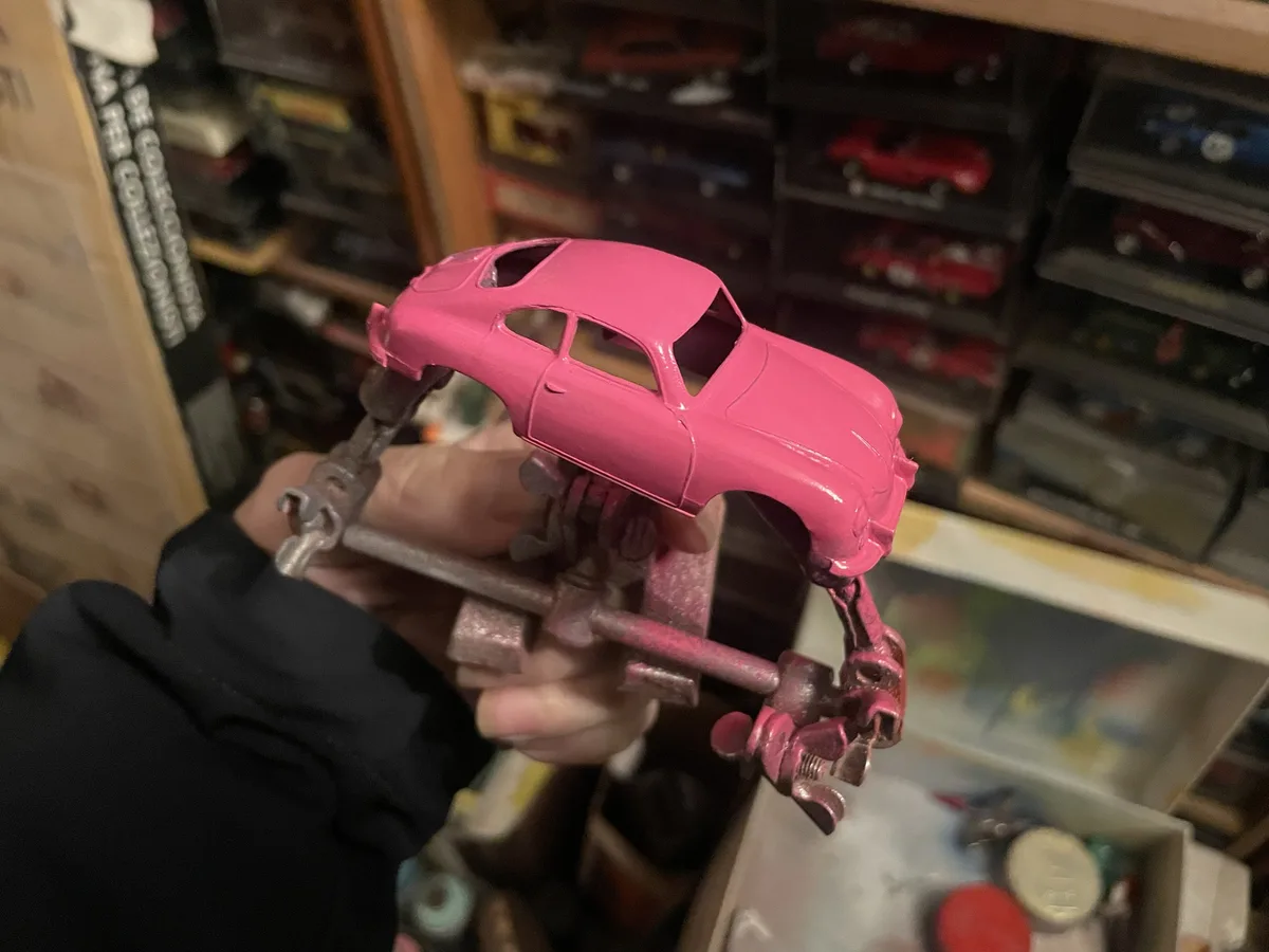 A hand holds a bright pink toy car body that is clamped to a rusty metal vise, with shelves of other model cars visible in…