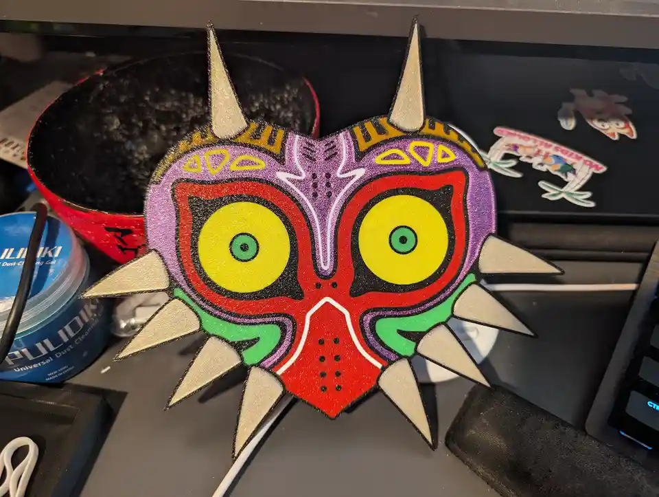 A colorful, glittery, 3D-printed replica of the Majora's Mask from The Legend of Zelda sits on a desk next to a keyboard, a…