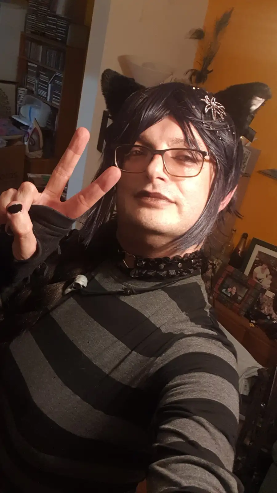A person with dark hair, glasses, and black cat ears and a spider hair accessory poses for a selfie, making a peace sign…