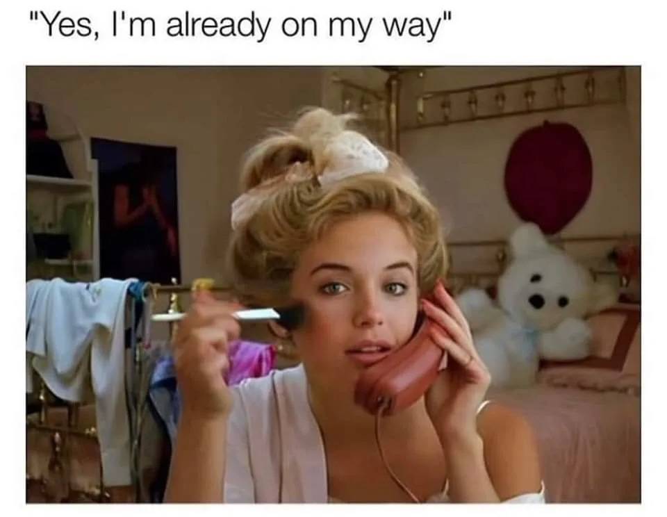 A woman with blonde hair in an updo, wearing a white robe, holds a red rotary phone to her ear while applying makeup with a…
