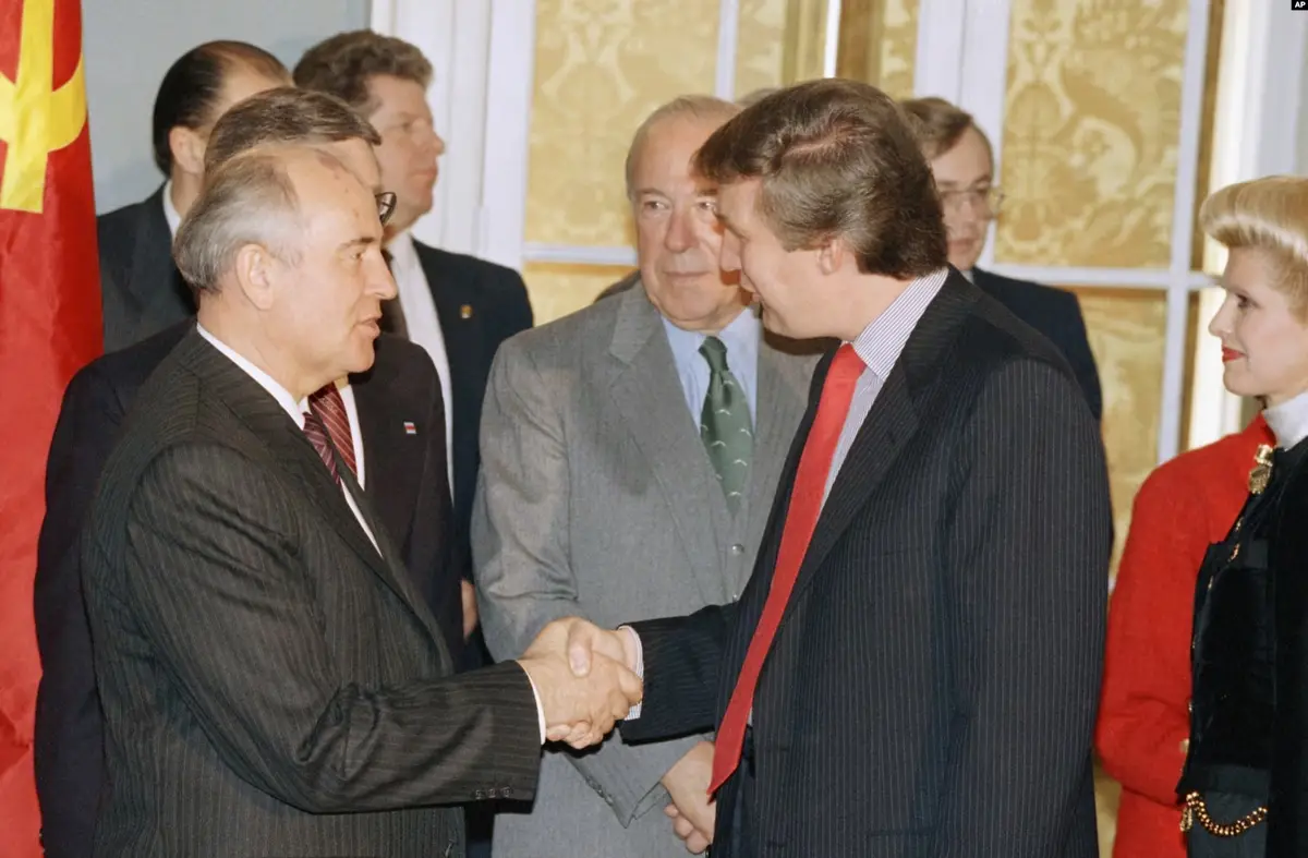 Two men in suits shake hands in a formal setting with a red flag featuring a hammer and sickle visible on the left. A woman…