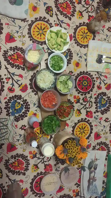 An overhead shot of a colorful, patterned tablecloth with various bowls of chopped ingredients for guacamole, including…