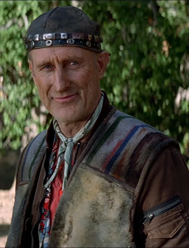 An older man with a weathered face, wearing a leather cap with metal studs, a striped vest over a patterned shirt, and a…