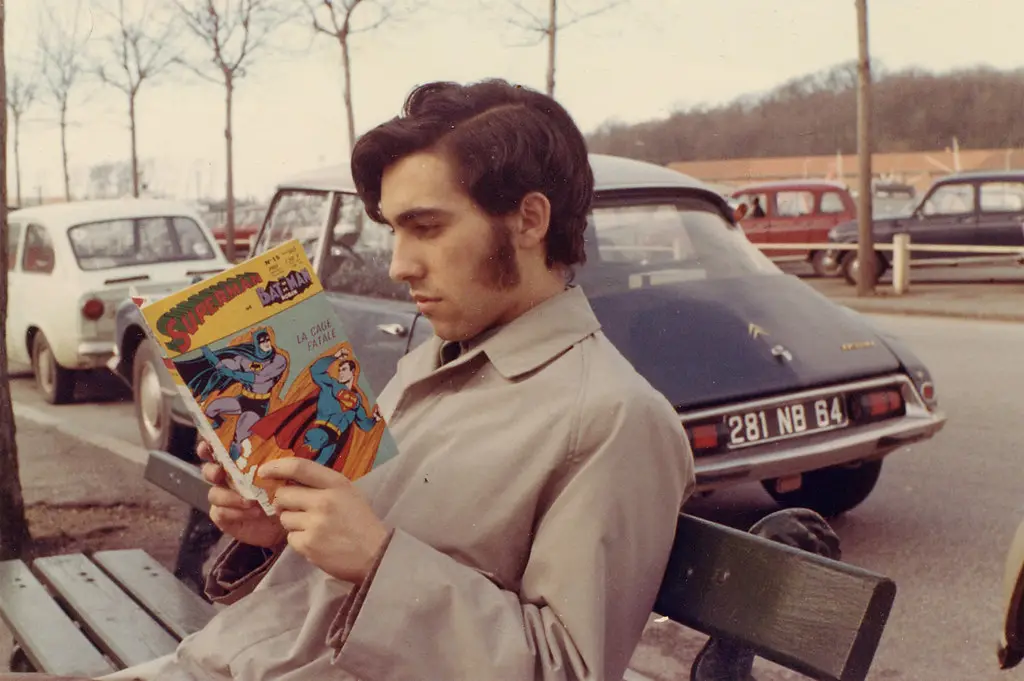 A man with dark hair and a light trench coat sits on a wooden bench outdoors, intently reading a vintage French comic book…