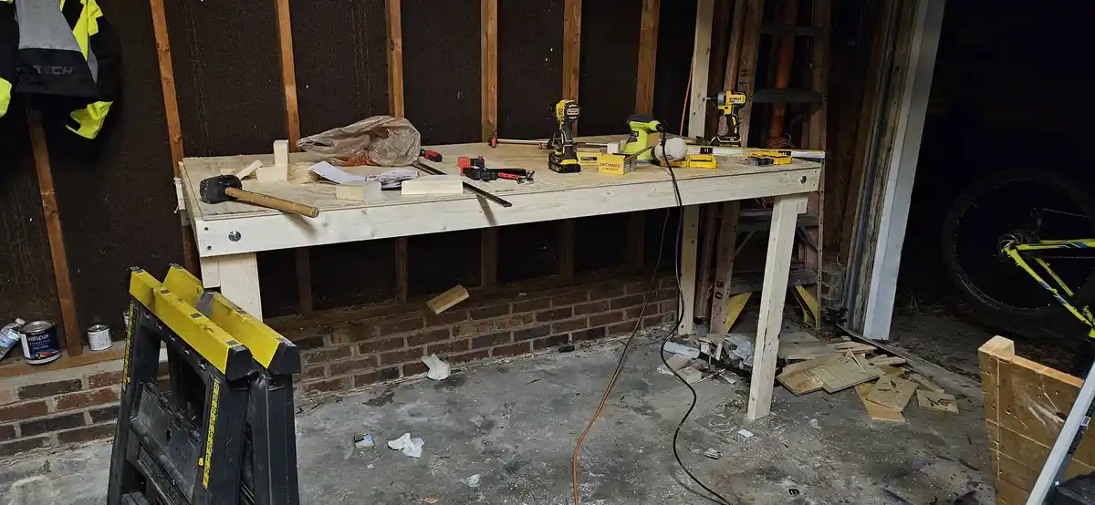 A cluttered garage workshop features a newly built wooden workbench with power tools, including DeWalt drills and a nail…
