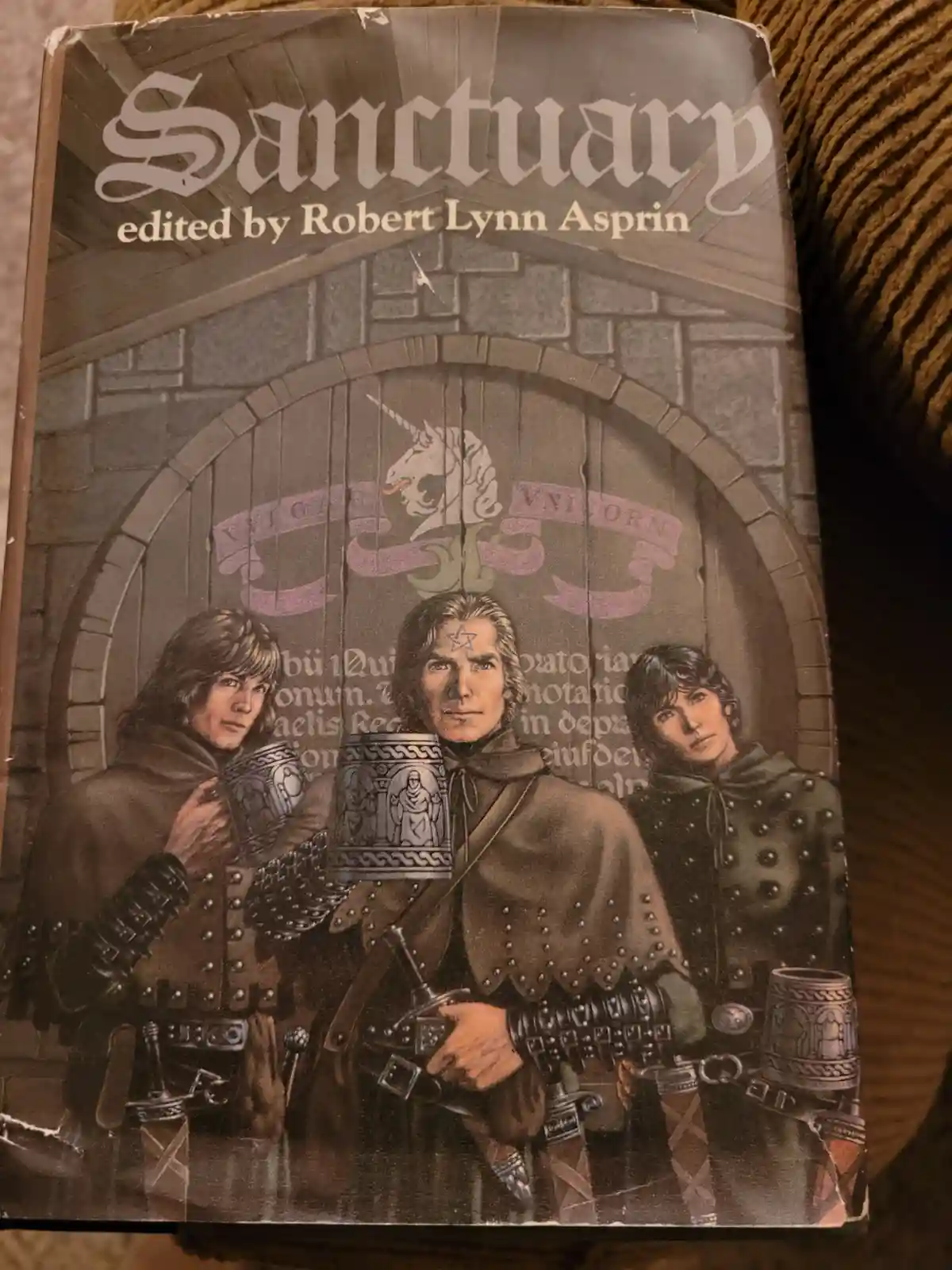 The image shows the cover of the book 'Sanctuary' edited by Robert Lynn Asprin, featuring a fantasy illustration of three…