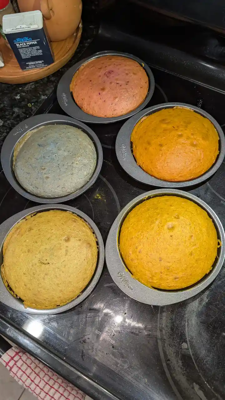 Five freshly baked cakes in metal pans sit on a black stovetop, displaying wildly varied colors: one pink, one blue, two…