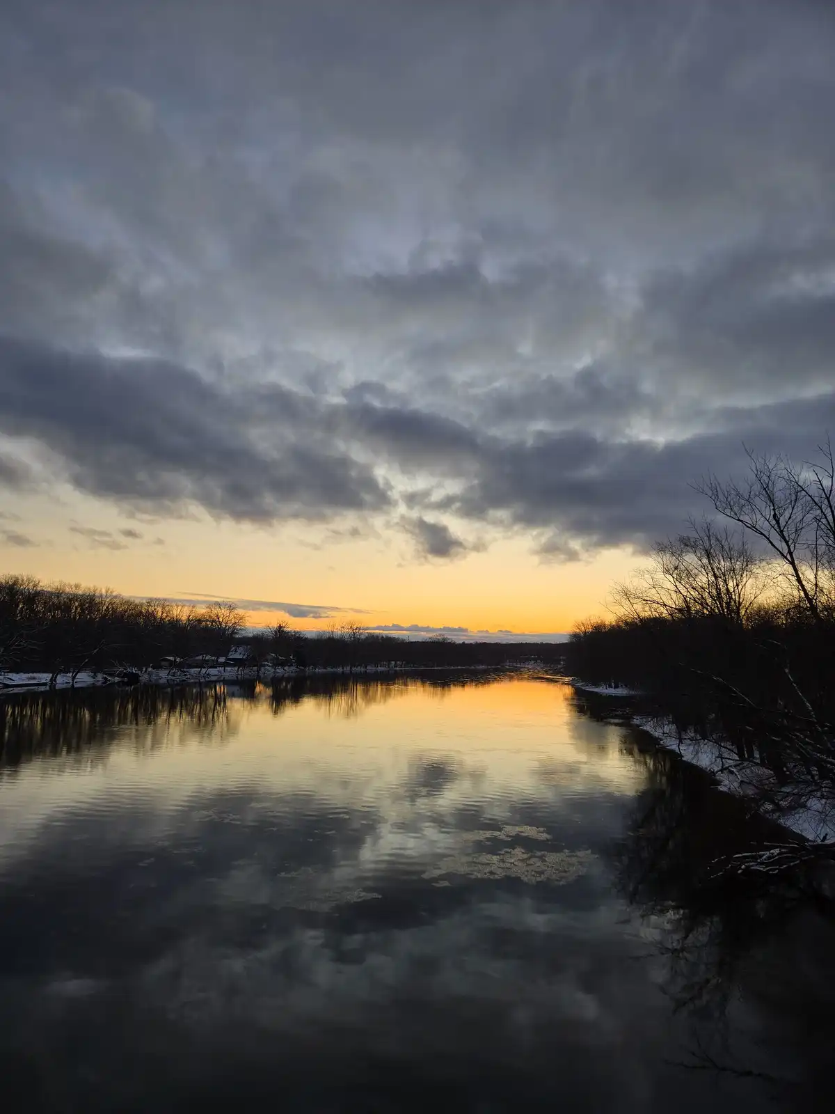 A serene winter sunset over a partially frozen river, with dark clouds above and bare trees lining the snowy banks…
