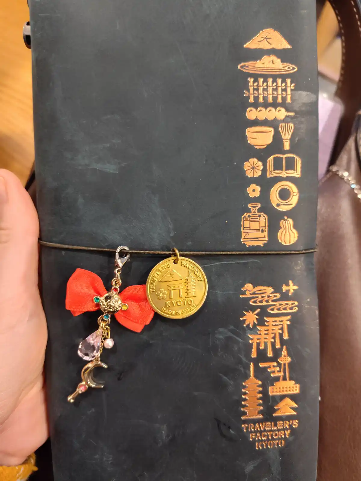 A hand holds a black leather travel journal from 'Traveler's Factory Kyoto' adorned with gold embossed Japanese icons like…