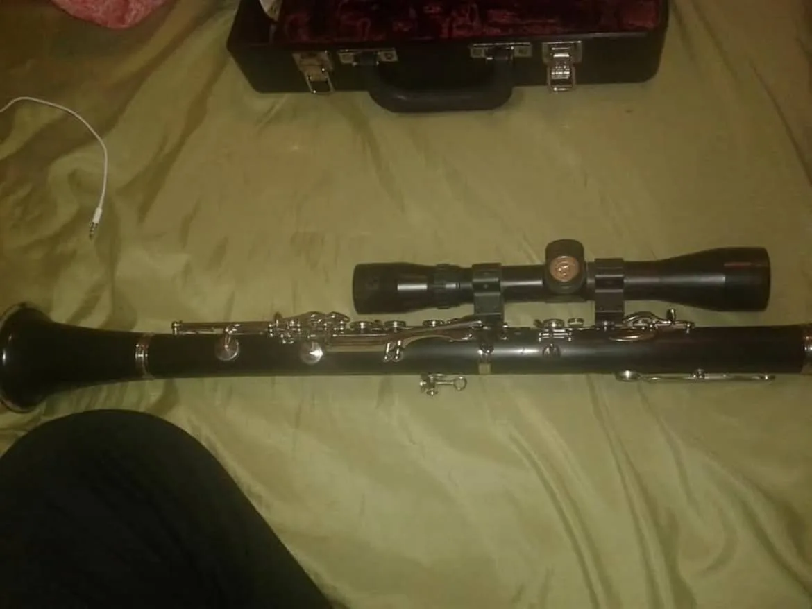 A clarinet lies horizontally on a green bedsheet, with a black rifle scope mounted on top of it. In the background, a black…