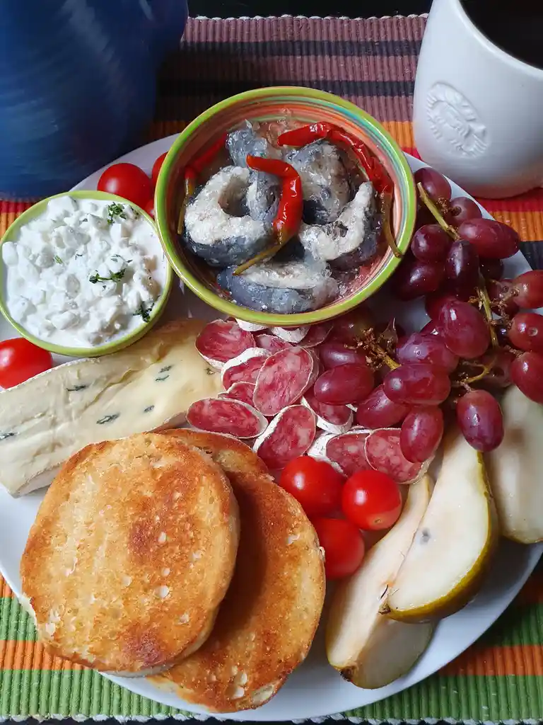 A plate of assorted foods including sliced salami, red grapes, cherry tomatoes, pear slices, toasted bread, blue cheese, a…