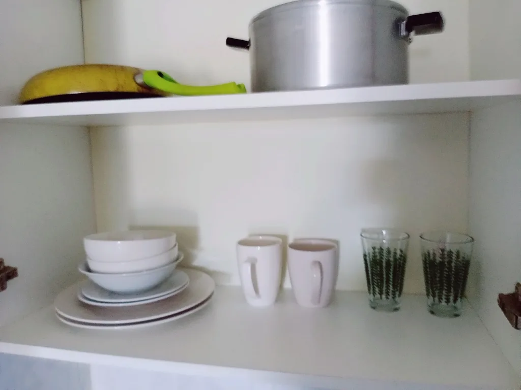 A kitchen cabinet shelf holds stacked white bowls and plates, two white mugs, and two patterned glasses. Above them, a…