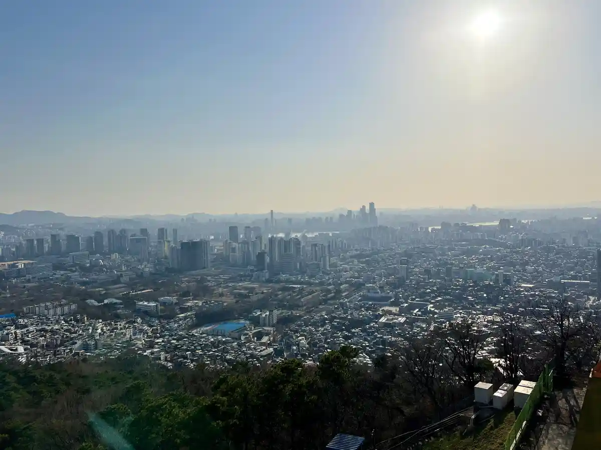 A wide-angle photo from a hilltop overlooking a sprawling, hazy cityscape with dense residential areas, modern skyscrapers…
