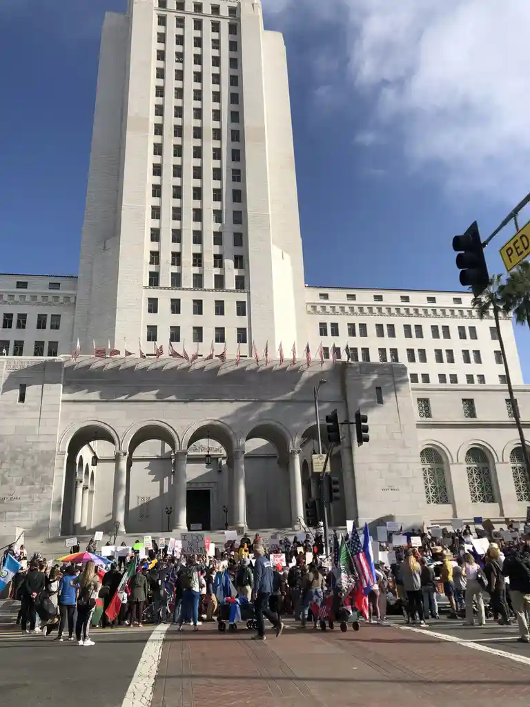 A large crowd of protesters holding signs and flags gathers in front of the tall, white Los Angeles City Hall building under…