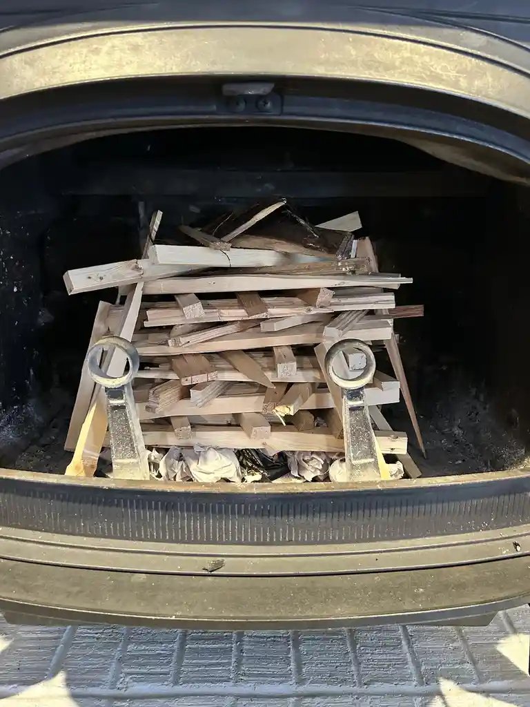 The image shows the open trunk of a car filled with a large pile of wooden planks and debris, with two metal fireplace tools…