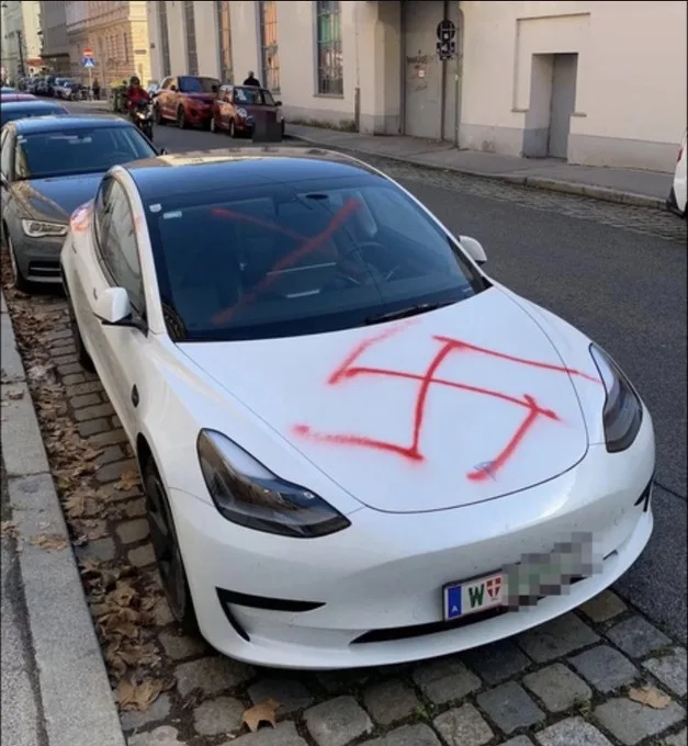 A white Tesla Model 3 parked on a cobblestone street has a large red swastika spray-painted across its hood and windshield…