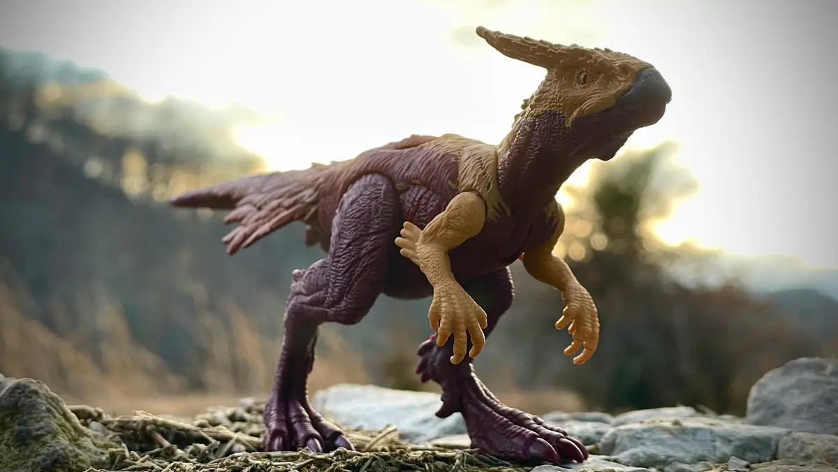 A plastic toy dinosaur figurine, resembling a theropod with a horned head and feathered body, stands on rocky terrain with a…