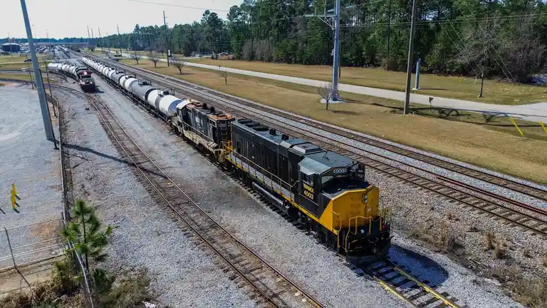 An aerial photo shows a long freight train with a black and yellow locomotive labeled 'WAMX 4003' pulling a line of white…