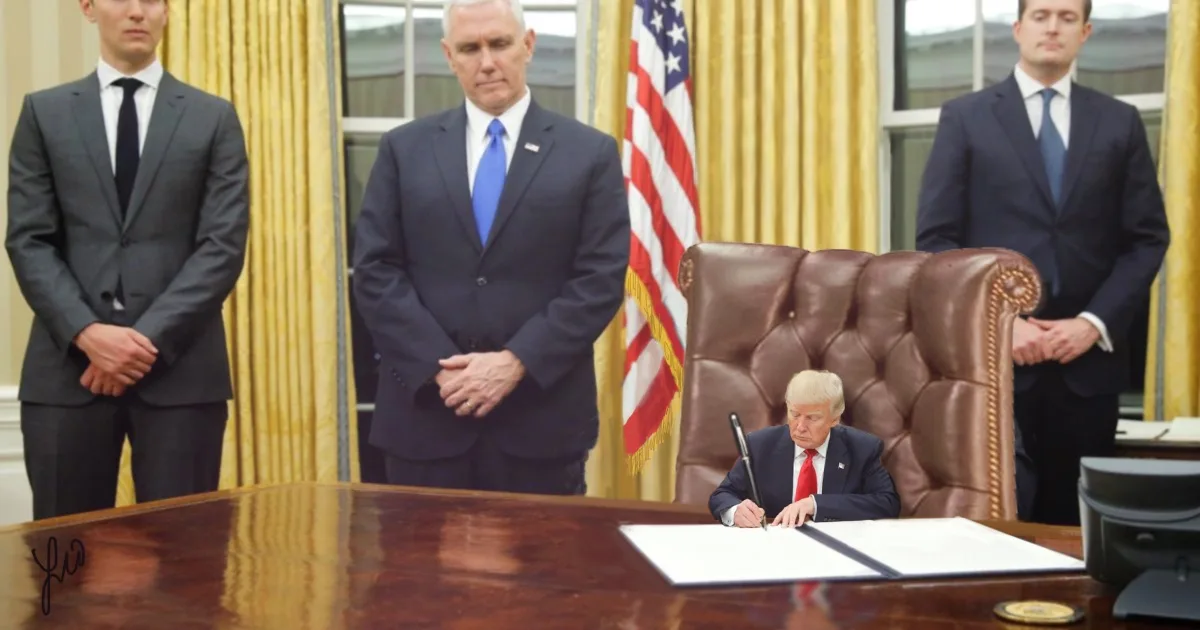 A man with blonde hair sits at a large wooden desk in an office with yellow curtains and an american flag, signing a…