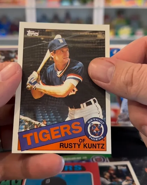 A hand holds a vintage Topps baseball card featuring Rusty Kuntz in a Detroit Tigers uniform, poised to bat. The card's text…
