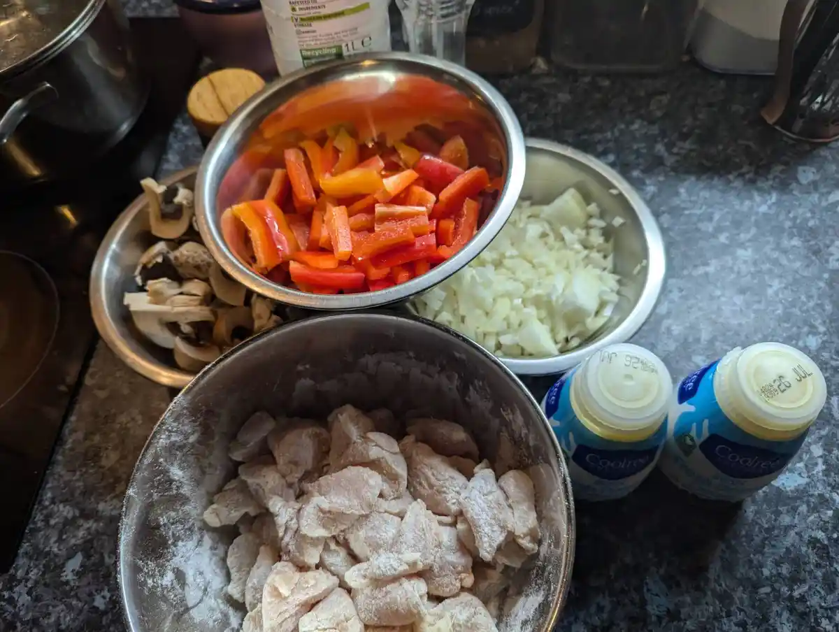 A kitchen counter displays three metal bowls: one with flour-dusted chicken pieces, one with chopped onions, and one with…