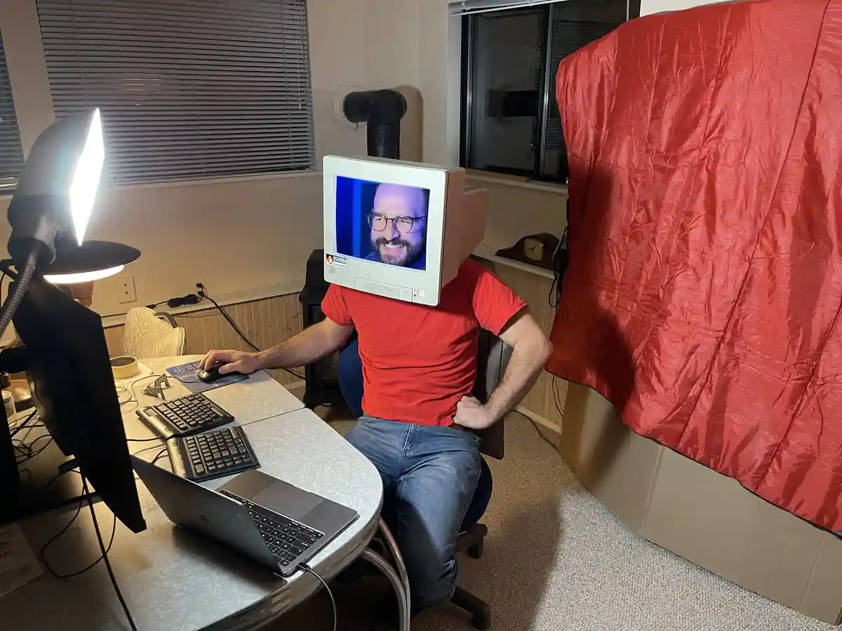 A person wearing a red shirt and jeans sits at a desk with a laptop and external keyboard, using a mouse. Their head is…