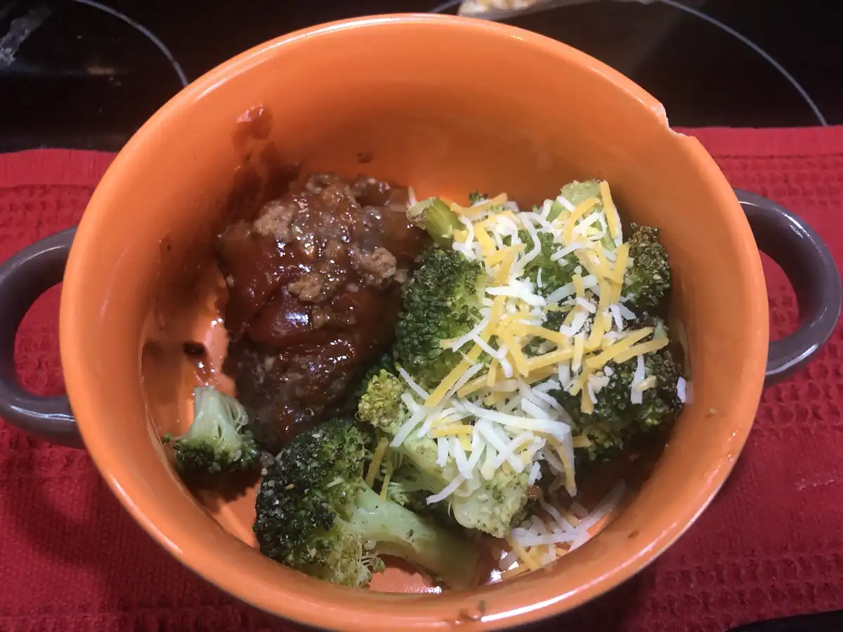 An orange ceramic bowl with handles sits on a red placemat, containing a serving of meatloaf in a brown sauce alongside…