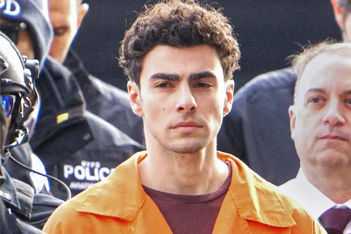 A man with curly dark hair wearing an orange prison jumpsuit stands solemnly, flanked by NYPD police officers and another…