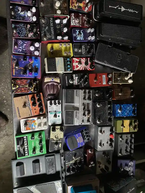 An overhead shot displays a densely packed pedalboard covered with dozens of colorful and varied guitar effects pedals, each…
