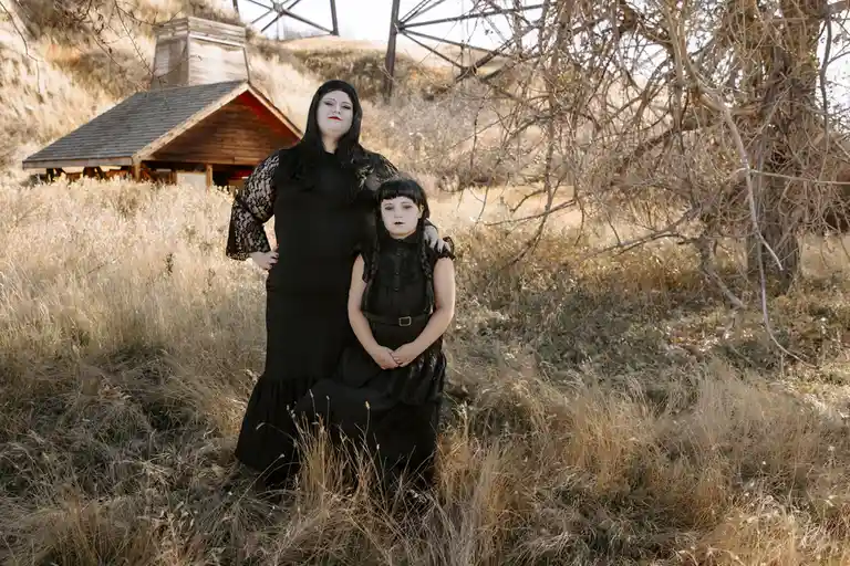 Two people dressed as Wednesday Addams and her mother, Morticia, pose in a field of dry grass with a wooden shed and a metal…