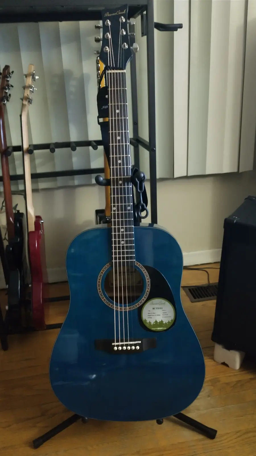 A blue acoustic guitar by 'Bourbon Creek' stands upright on a stand in a room with hardwood floors and other guitars on a…