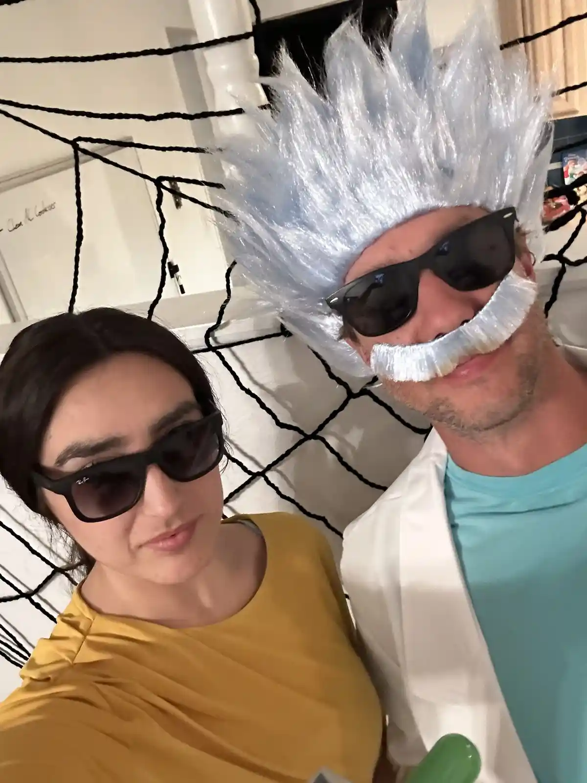 A selfie of two people, one dressed as a scientist with a spiky white wig, sunglasses, and a matching white mustache…