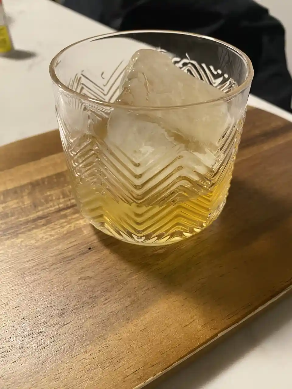 A close-up photo shows a glass with a large ice cube and amber-colored liquid, likely whiskey, resting on a wooden tray. The…