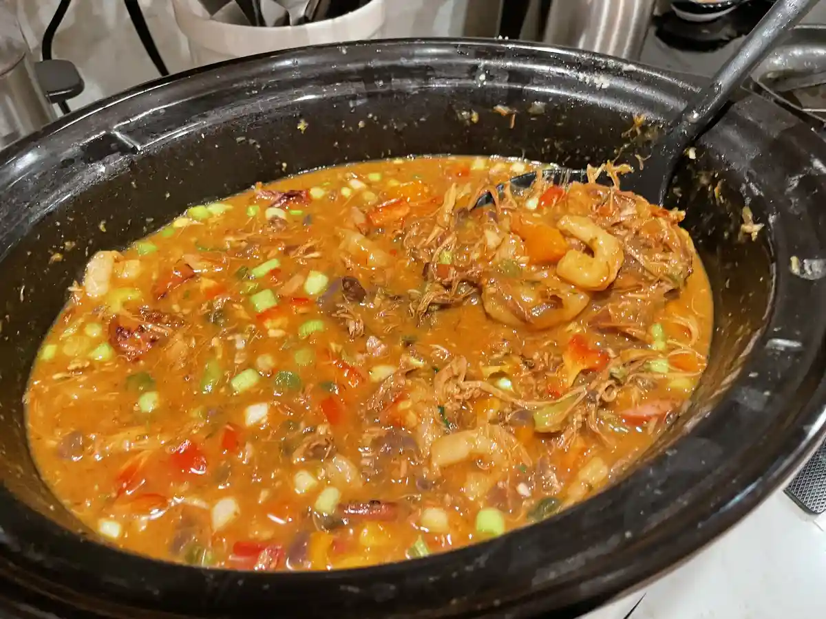 A close-up photo shows a black slow cooker filled with a thick, orange-brown stew containing shredded meat, chopped green…