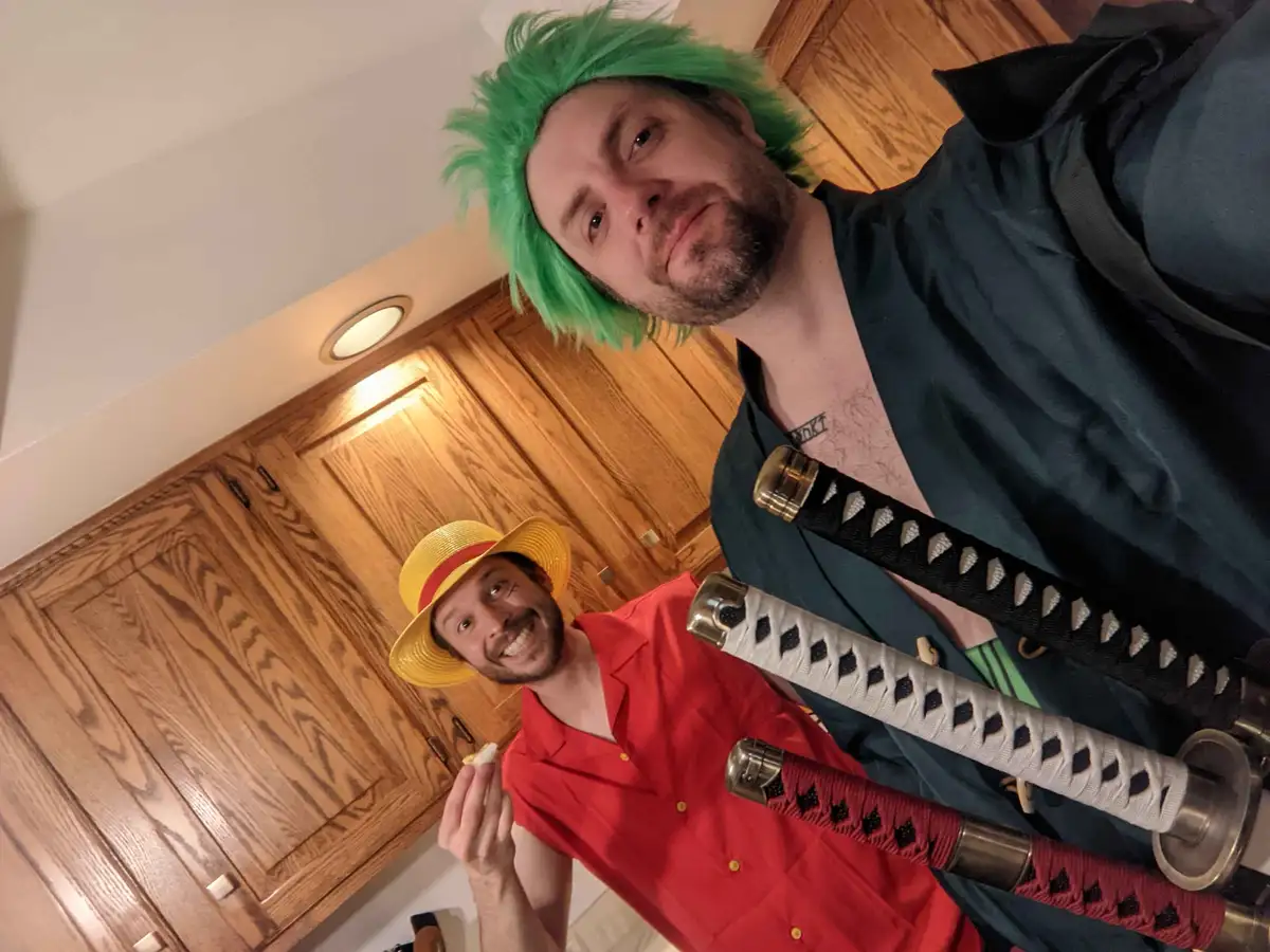 Two men in a kitchen, one with green wig and swords (Zoro cosplay) looks smugly at the camera, while the other in red shirt…