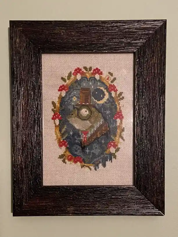 A framed cross-stitch artwork depicts a whimsical, anthropomorphic frog wearing a top hat and monocle, set against a night…