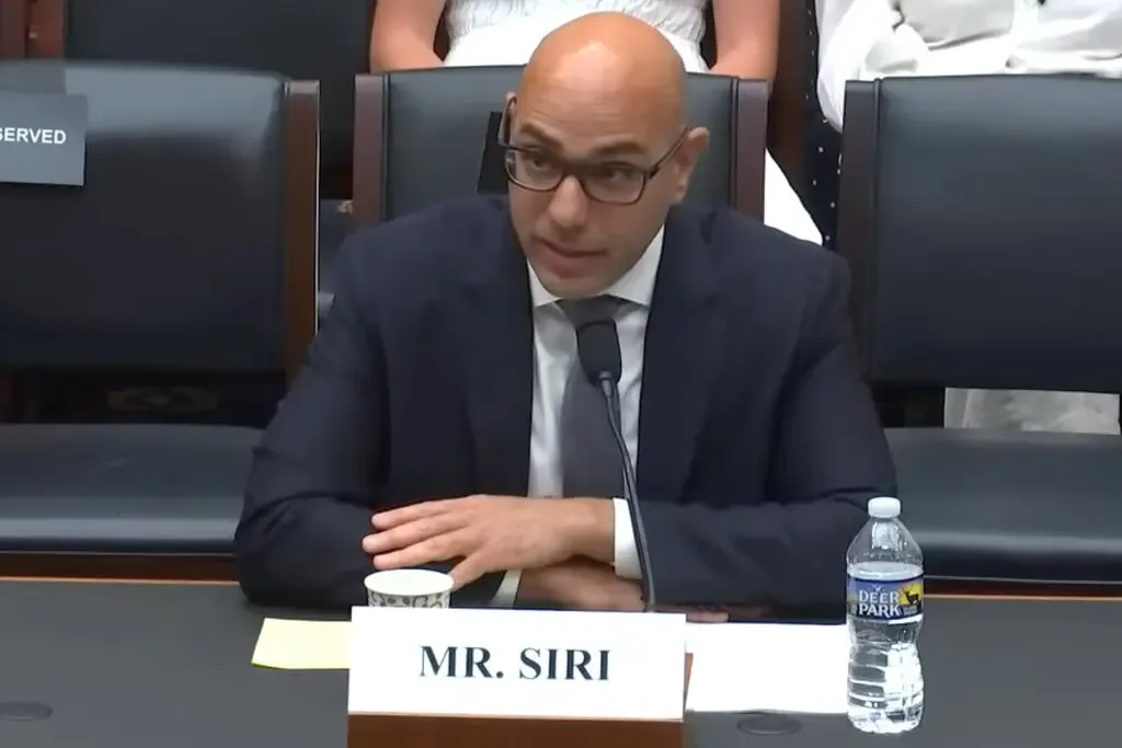 A bald man wearing glasses and a suit sits at a formal hearing table with a nameplate reading 'MR. SIRI' in front of him. He…