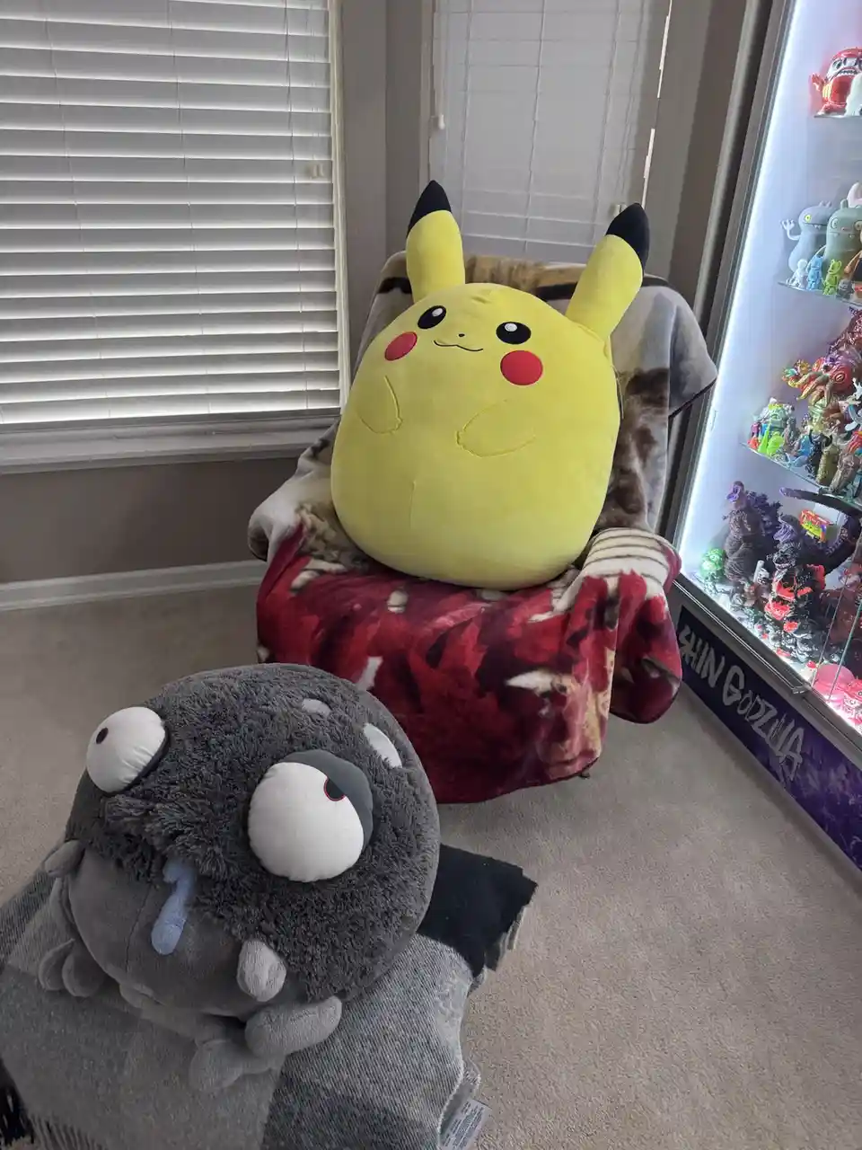 A large yellow Pikachu plush toy is seated comfortably in a recliner chair covered with blankets, looking relaxed. In the…
