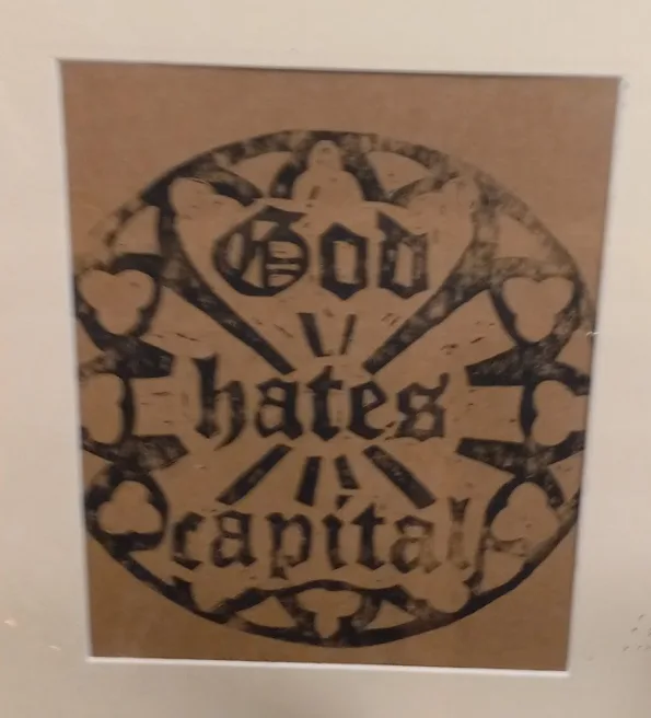 A framed, aged-looking print on brown paper displays the text 'God hates capital' in a stylized, gothic font, arranged…