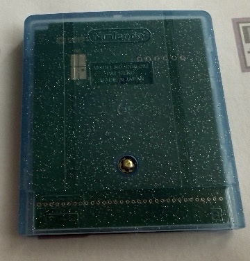 A close-up photo of a green Nintendo Game Boy cartridge with a translucent blue plastic shell, showing the cartridge's metal…