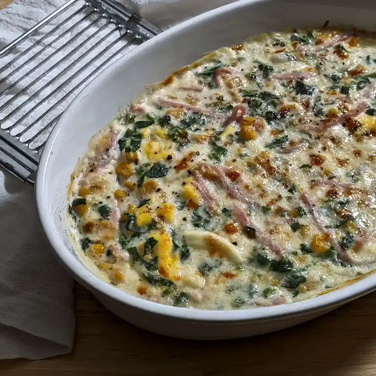 A close-up photo of a freshly baked casserole in a white oval baking dish, featuring melted cheese, green leafy vegetables…