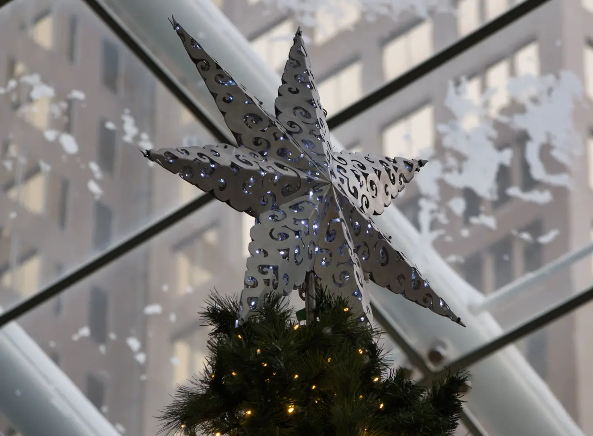 A close-up photo shows a large, ornate, silver star ornament with intricate scrollwork and embedded lights, perched atop a…