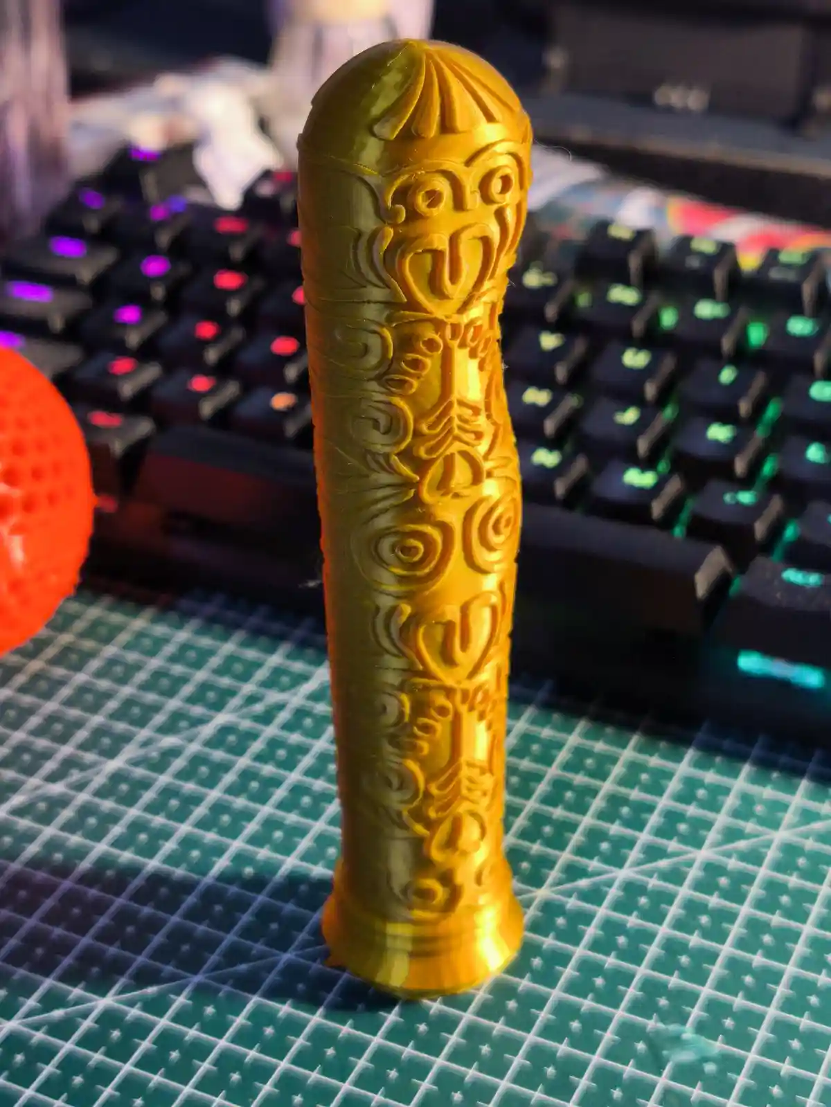A golden, intricately carved cylindrical object resembling a stylized phallus with tribal patterns stands upright on a green…